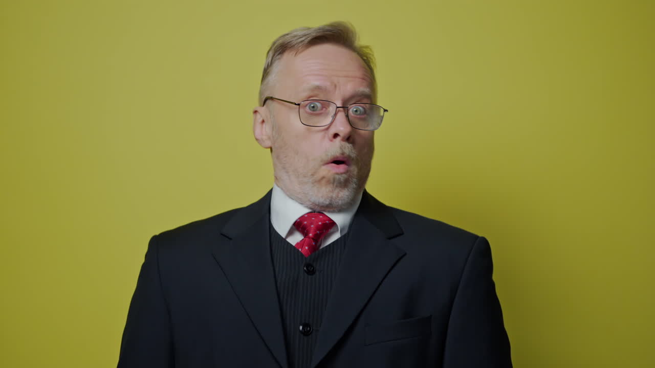 Positive businessman showing different emotions. Mature man in glasses and elegant costume shows funny face on the camera on yellow studio background.