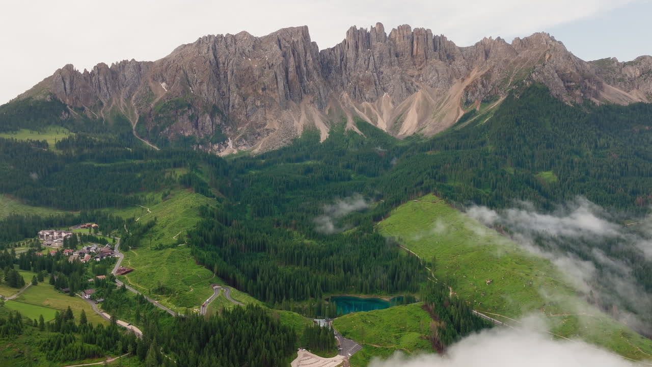 Slow panning drone shot of Lago di Carezza in the Dolomites, Italy. Emerald-green waters surrounded by spring forests and dramatic alpine peaks in the distance