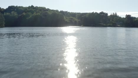 aerial view flying low over rippling lake water surface towards sun glare and forested shore, marcillac la croisille, france