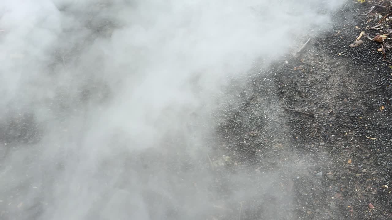 Dense white smoke drifts over gravel surface, natural daylight, static overhead camera, outdoor environment