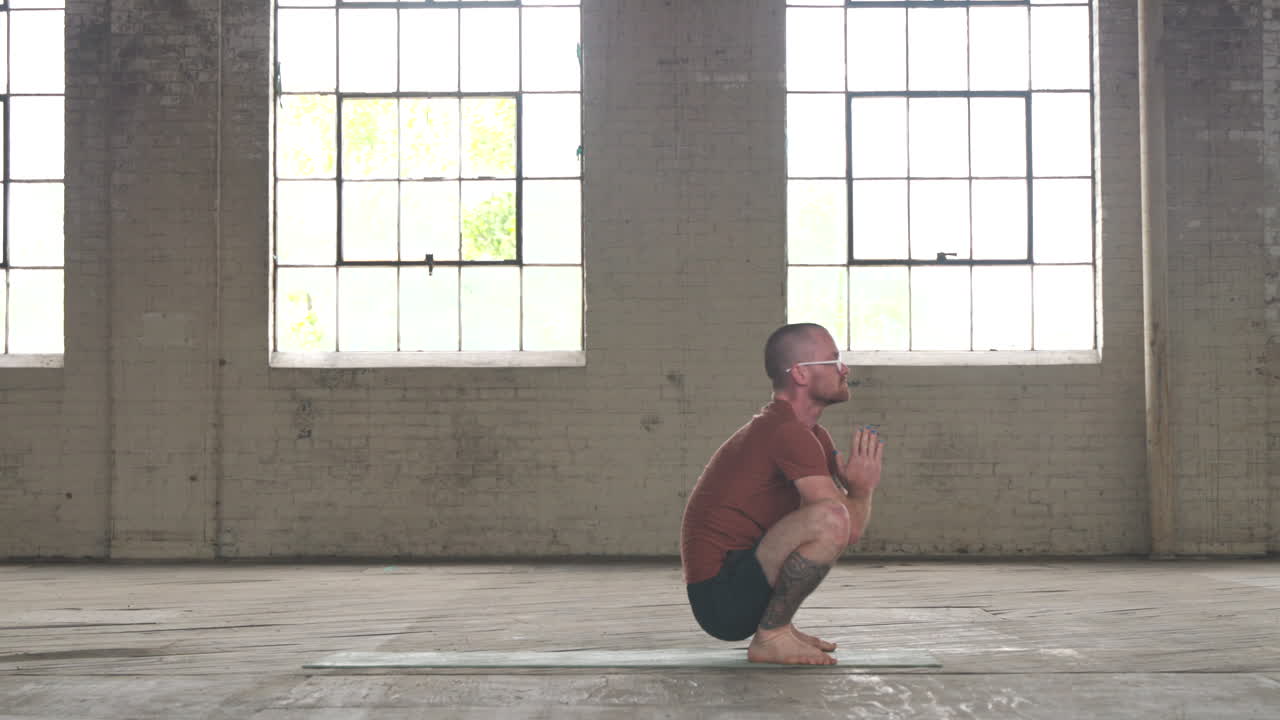 Man in an industrial warehouse practicing yoga doing a malasana squat.