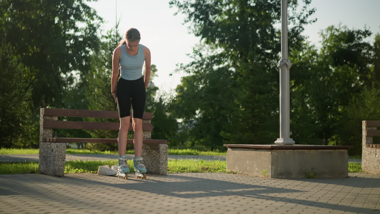 una mujer joven vestida con una camiseta azul claro y medias negras se levanta de un banco en el parque mientras se equilibra en patines, con zapatillas blancas colocadas debajo del banco, rodeada de vegetación