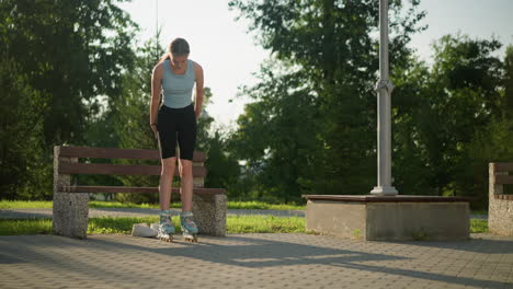 young woman wearing light blue top and black tights stands up from bench in park while balancing on roller skates, with white sneakers placed under bench, surrounded by greenery