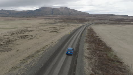 Vista-Aérea-Con-Seguimiento-Desde-Detrás-De-Un-Automóvil-Azul-Y-Sobre-El-Camino-De-Tierra-Que-Conduce-A-La-Formación-Rocosa-Islandesa-De-Hvitserkur-En-Un-Día-Aburrido