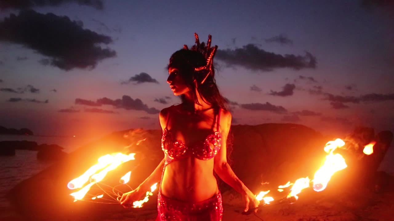 Erotic and sensual belly dancer performing spinning and figure 8 moves with fire props on the beach during golden hour.
