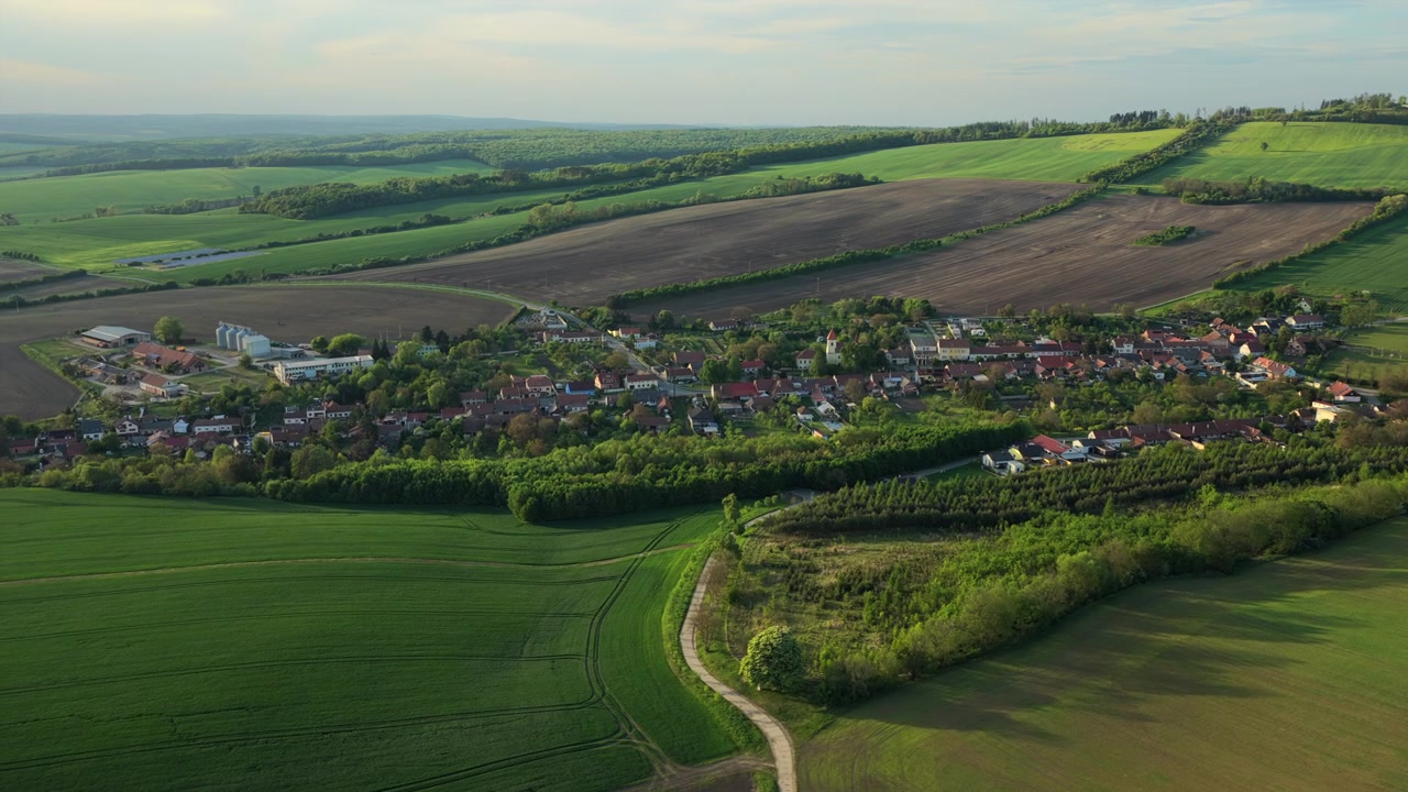 Dronebilde av en sprøytetraktor på et grønt åkerfelt i åssiden med utsikt mot bølgende skogkledde åser i varmt kveldslys, Moravia, Tsjekkia