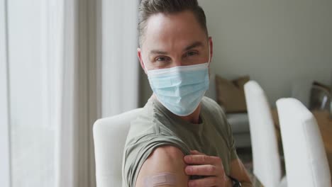 Portrait-of-caucasian-man-wearing-face-mask-showing-his-vaccinated-shoulder-at-home