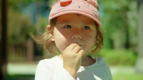 Gesicht-Eines-Blonden-Zweijährigen-Mädchens,-Das-Weiße-Vitaminpillen-Lutscht-Und-Langsam-Isst,-Während-Es-In-Einem-Park-Steht-Und-Tagsüber-In-Die-Kamera-Schaut