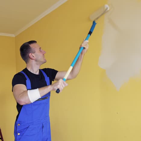 room improvement with a new color. professional worker in blue overalls painting wall with a paint roller on a long handle on. craftsman working