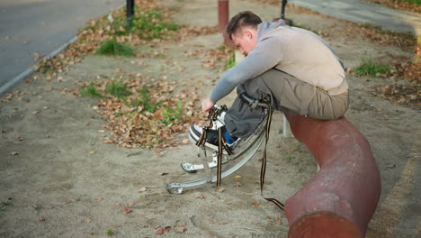 man sitting on curved log in park, wearing light gray sweatshirt and olive-green pants, focused on adjusting straps of spring stilts with a determined expression, surrounded by fallen leaves