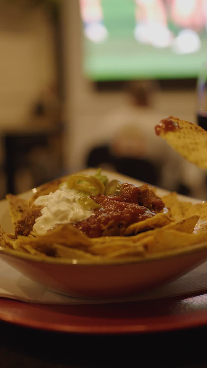 Mano che intinge un chip di mais in salsa e crema acida, in un ambiente da bar, con una partita sportiva in televisione sullo sfondo.
