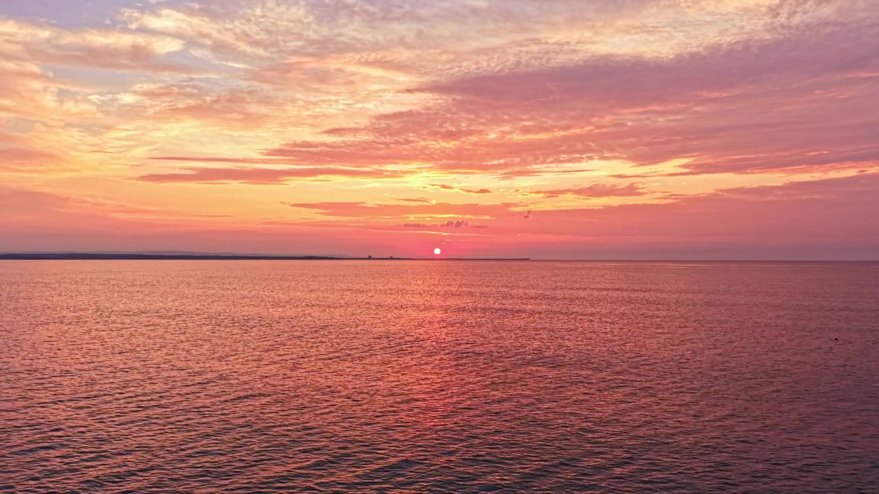 Bright sunset over the water reflects shades of orange and pink with clouds in the sky and calm waves in the background
