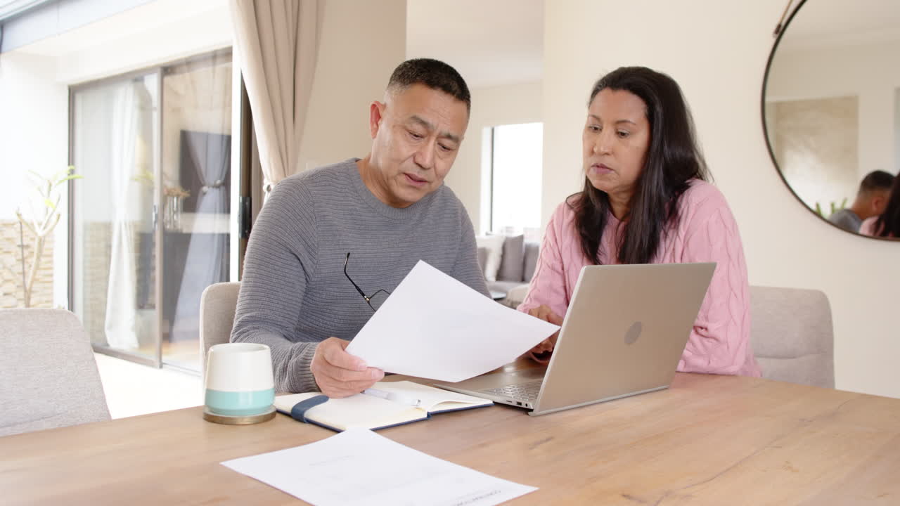 Senior asian couple reviewing documents together at home using laptop, focused and engaged