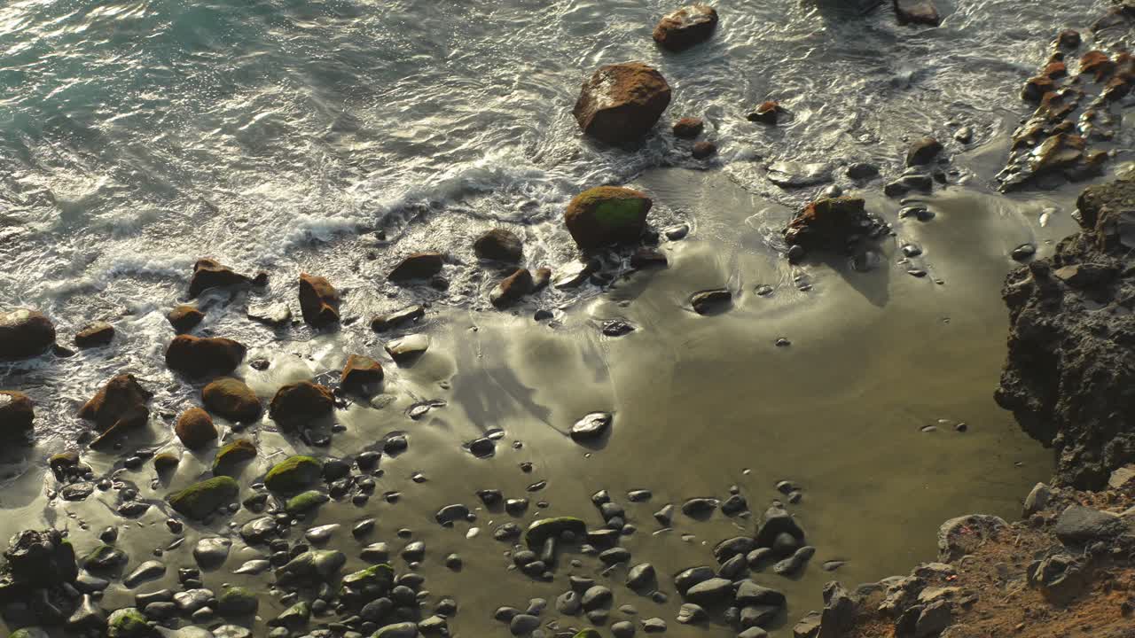 Optagelse fra oven af bølger der slår ind mod en stenet sort sandstrand