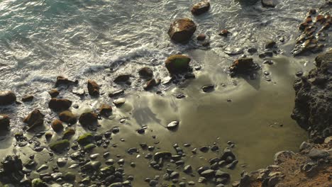 Top-down-shot-of-waves-crashing-into-rocky,-black-sand-beach