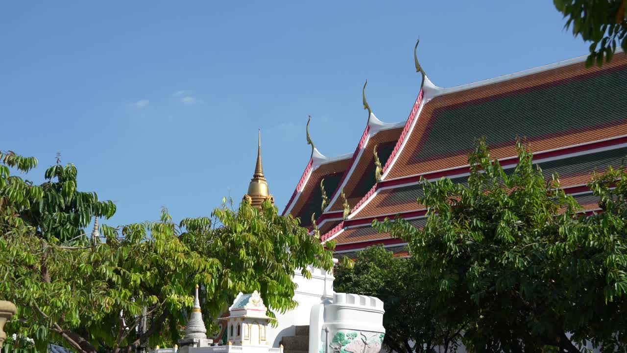 Exterior view of Wat Pho, Bangkok, featuring the gilded Chofa finials and tiered, tiled temple roof, with a smaller golden chedi (stupa) and lush trees beneath a clear blue sky