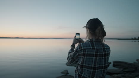 woman photographing sunset over lake with smartphone