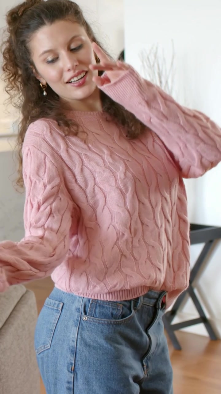 Vertikal video: Kvinde holder telefon, udfører flydende kropsbevægelser i hjemmet, lyserød strikket top, jeans, beige sofa.