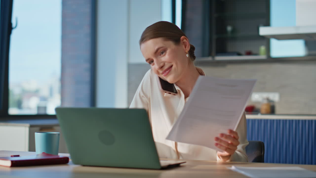 Professional woman talking smartphone working at laptop and handling documents