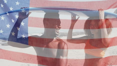 american flag waving against african american couple carrying surfboard on the beach