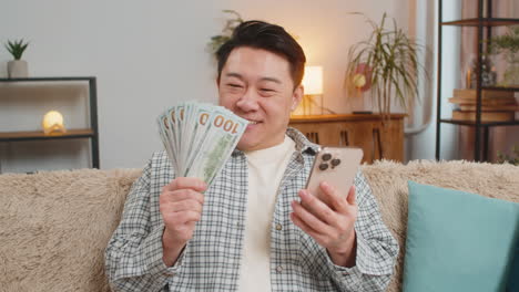 successful middleaged man holding smartphone and dollar bill money fan sitting on couch at home