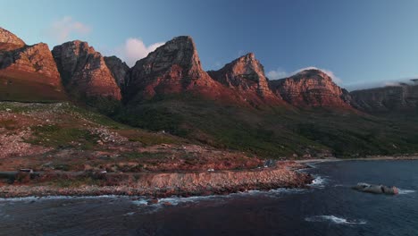 Victoria-Road-Con-Las-Montañas-De-Los-Doce-Apóstoles-En-Ciudad-Del-Cabo,-Sudáfrica---Toma-Aérea-De-Un-Drone