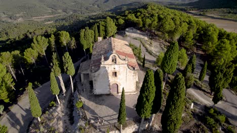 Backwards-aerial-revealing-top-view-of-an-aisled-Hermitage-on-top-of-a-hill-among-trees-in-Spain