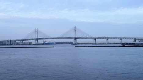 Distant-view-of-a-suspension-bridge-over-serene-water-with-boats,-under-a-cloudy-sky