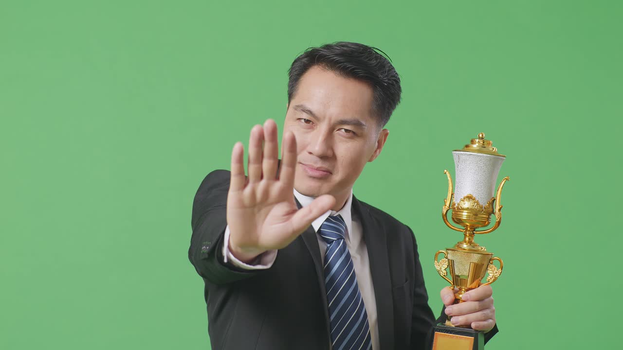 슈트와 타이를 입은 아시아 사업가, 금메달과 트로피를 들고, 손짓을 하지 않고 카메라를 향해 미소 짓고, 녹색 스크린을 배경으로 스튜디오에서