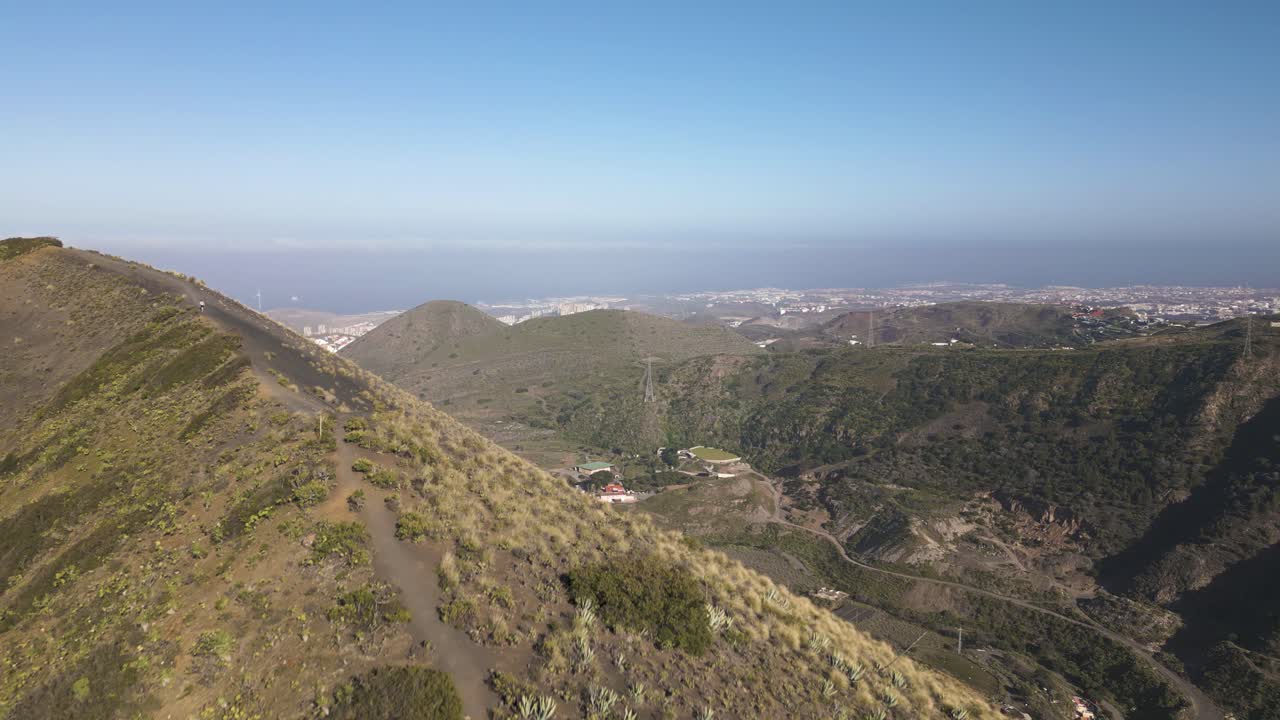 vuelo aéreo hacia adelante sobre el parque natural de la caldera de bandama con personas de senderismo en el camino en la cima de la montaña - océano y ciudad en el fondo - gran canaria, islas canarias durante un hermoso día soleado
