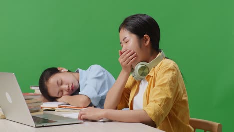 close up of asian woman student writing and yawning while her friend is sleeping on a table with a laptop in the green screen background classroom