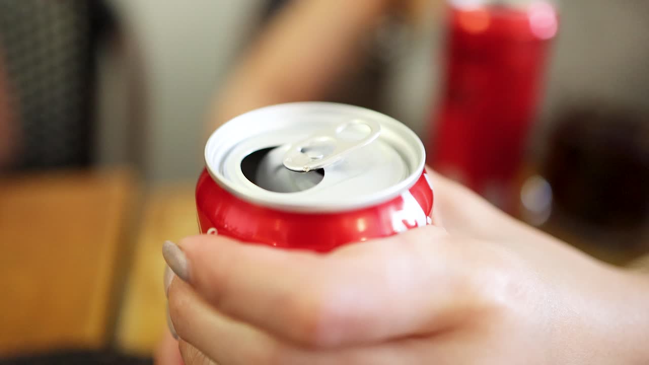 A close-up sequence shows a person’s hand opening a red soda can on a wooden table, with soft natural lighting and shallow depth of field