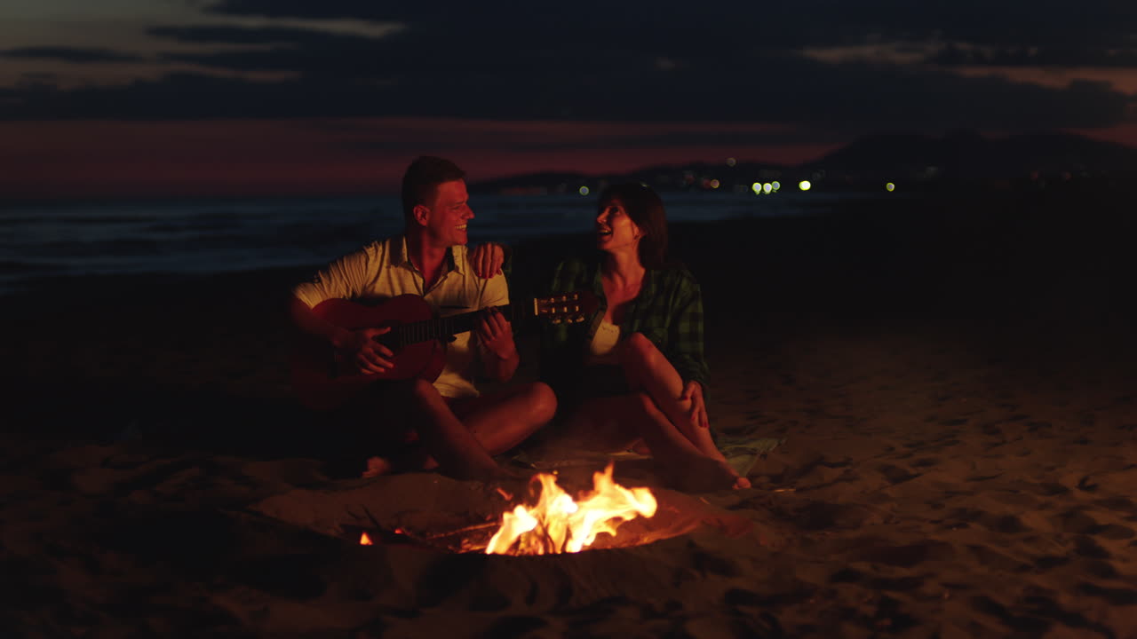 pareja tocando la guitarra junto a una fogata en la playa por la noche