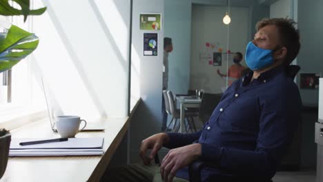 Thoughtful-caucasian-man-wearing-face-mask-using-laptop-and-looking-out-of-window-at-modern-office