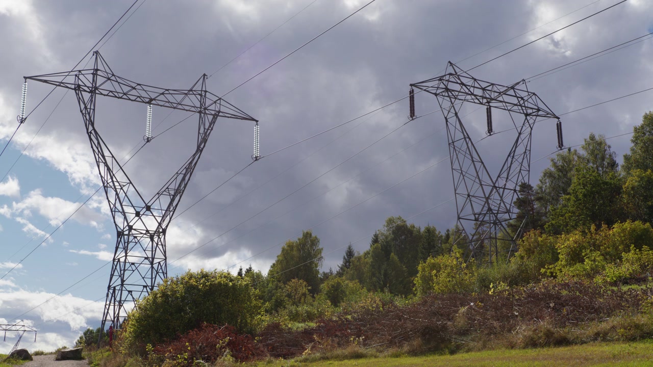 Steel power pylons carry high voltage lines over a cut woodland edge with dense trees and moving clouds in Norway, useful for energy or infrastructure themes.