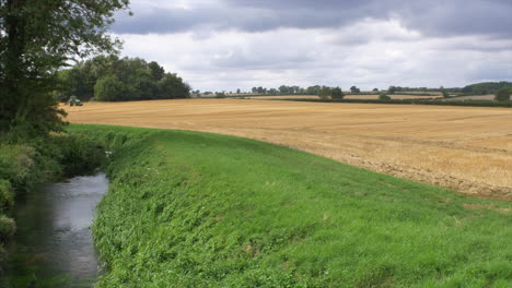 Tractor-making-a-circular-movement-in-the-distance-in-a-beautiful-field,-with-a-running-stream-in-the-foreground