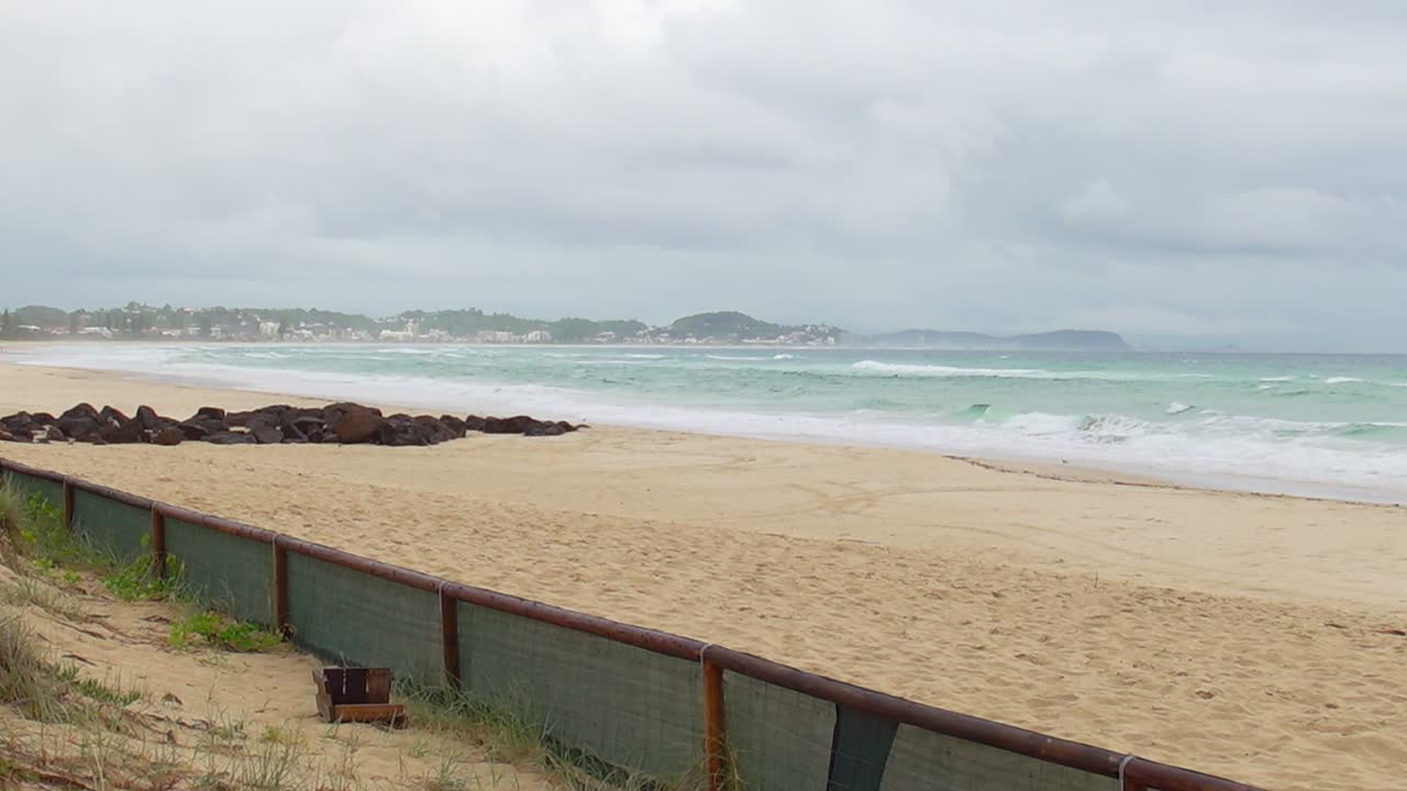 Waves crash along North Kirra Beach as sand dunes and protective fencing line the coast under a clear blue sky.