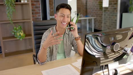 happy asian male creative talking on smartphone using computer at desk in casual office, slow motion