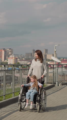 woman walks on viewpoint with daughter sitting in wheelchair. mother and preschooler girl with cerebral palsy enjoy scenic city view on sunny spring day