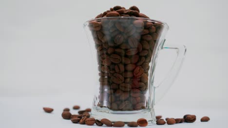 Foto-De-Estudio:-Granos-De-Café-Dentro-Y-Alrededor-De-Una-Taza-De-Café-Alta-Y-Transparente-Con-Asa