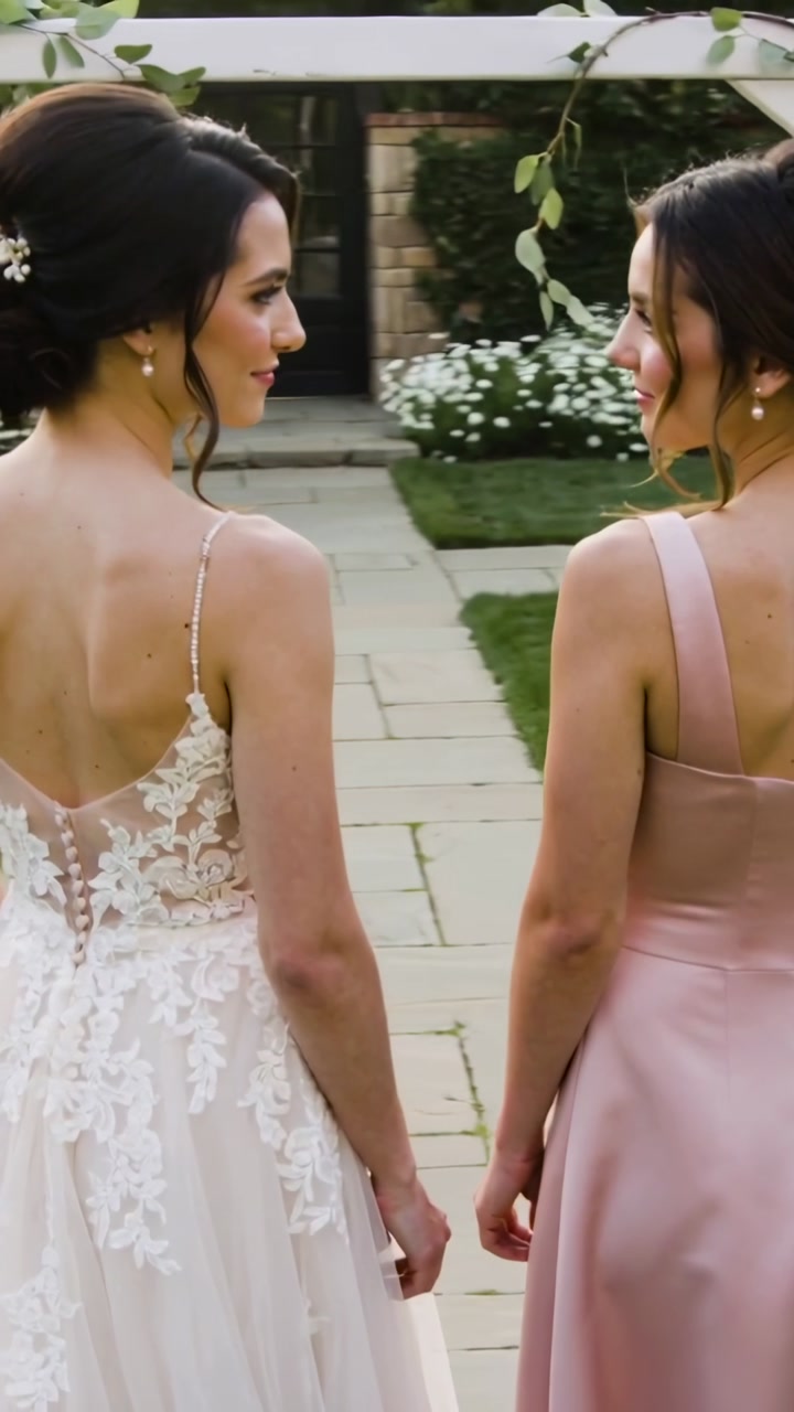 Bride in lace and bridesmaid in blush beneath arbor at wedding, starting head turns before ceremony