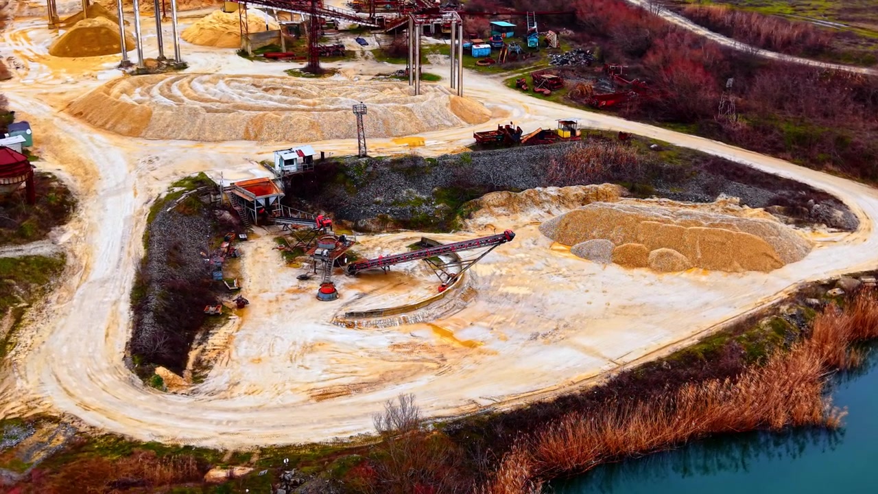 Aerial view of gravel processing site by lake, Bulgaria. Wide angle shows conveyors, sand mounds, a dry reed bed, and distant water reflections.