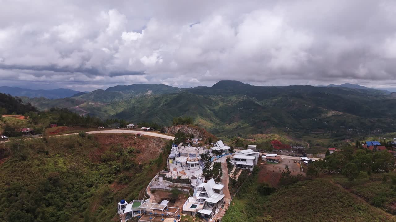 Scenic 4K aerial drone shot of a white architectural complex (resort or landmark) situated on a steep mountain peak. Overlooking deep green valleys and winding roads under a cloudy sky