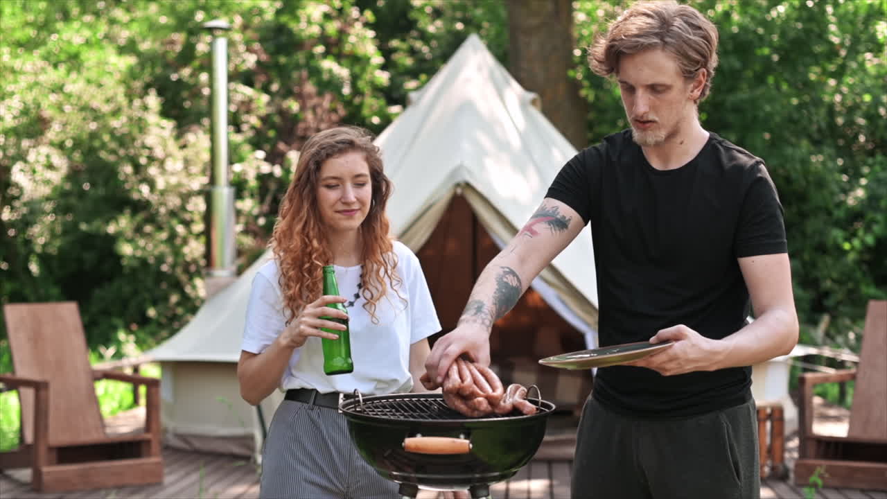 Young couple frying meat on the grill and drinking beer. Greenery around. Glamping. Slow motion