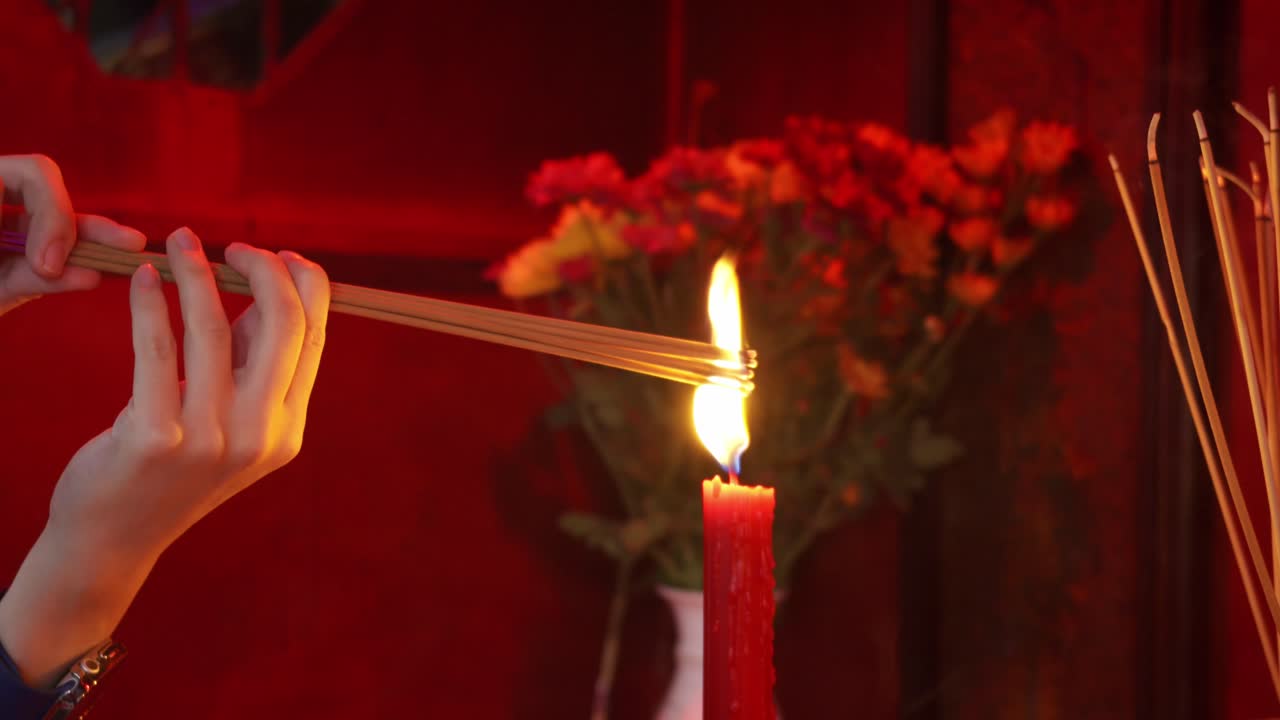 A person lights incense sticks from a red candle flame during a traditional ritual, with flowers and offerings in the background, symbolizing prayer, respect, and spirituality