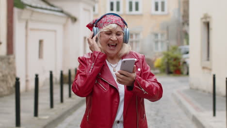 happy senior woman in wireless headphones choosing listening music in smartphone dancing outdoors