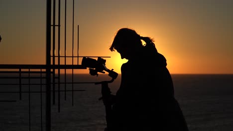 A-female-filmmaker-recording-video-with-a-camera,-silhouetted-by-a-beautiful-sunset