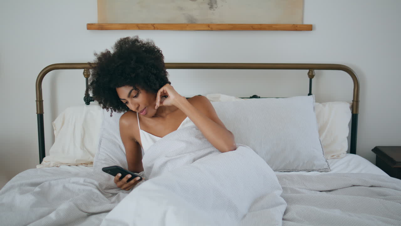 Ragazza che si sveglia sbadigliando a letto al mattino. Donna africana che legge al telefono sdraiata sul cuscino.
