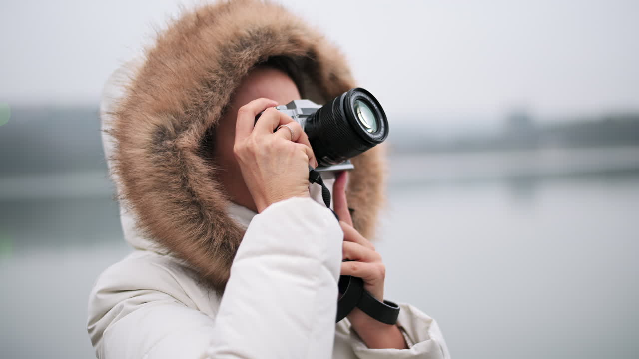 A woman wearing a warm coat with fur hood captures beautiful moments by the water's edge. The backdrop features soft gray skies on a chilly day, creating a serene atmosphere