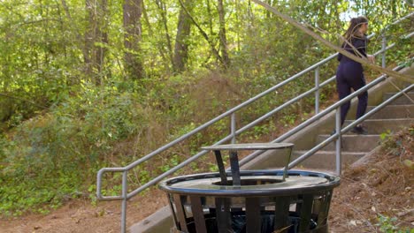 a young woman tosses garbage into a trashcan on her way up a large flight of stairs in a wooded park